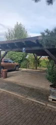Ein überdachter Parkplatz mit einem Auto und Bäumen im Hintergrund. Der Boden ist gepflastert und es gibt einen Müllbehälter daneben.