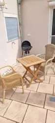 Eine kleine Terrasse mit einem Holztisch und zwei Stühlen. Im Hintergrund steht ein Grill.