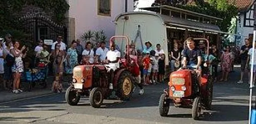 Zwei rote Traktoren fahren durch eine Straße, während eine Menge von Zuschauern zusieht. Es ist eine festliche Atmosphäre mit Menschen jeden Alters.