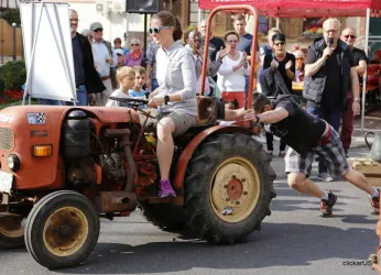 Eine Frau sitzt auf einem Traktor, während ein Mann ihn festzieht. Zuschauer beobachten die ungewöhnliche Aktion auf einem belebten Platz.