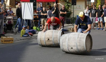 Zwei Männer rollen Fässer auf einer Straße, während eine Zuschauergruppe zusieht. Die Veranstaltung scheint ein Wettkampf oder ein Fest zu sein.