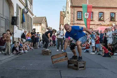 Eine lebhafte Straßenveranstaltung mit Menschen, die auf Holzkisten stehen und umgeben von Zuschauern sind. Kinder und Erwachsene beobachten begeistert die Darbietung.
