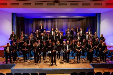 Ein großes Orchester steht auf der Bühne, angeordnet in zwei Reihen. Die Musiker tragen formelle Kleidung und lächeln in die Kamera.