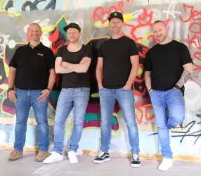 Vier Männer stehen lässig vor einer farbenfrohen Wand mit Graffiti. Sie tragen schwarze T-Shirts und Jeans, und ihre Körperhaltungen sind entspannt.