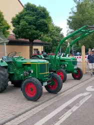 Zwei grüne Traktoren stehen auf einer Straße, umgeben von Bäumen. Ein Mann befindet sich in der Nähe und betrachtet die Traktoren.