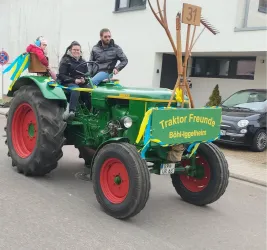Ein grüner Traktor mit roten Reifen fährt eine Straße entlang. Darauf sitzen zwei Personen und ein Kind, mit einem Schild, das "Traktor Freunde" zeigt.