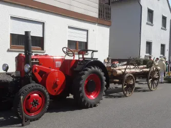 Ein roter Traktor steht auf der Straße neben einem Wagen mit Holzstämmen. Im Hintergrund sind Wohnhäuser zu sehen.