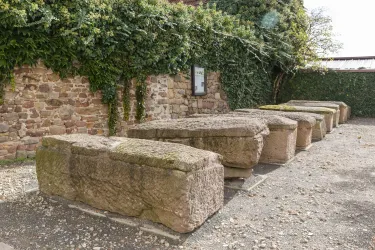Eine Reihe von großen, steinernen Blöcken steht vor einer Mauer mit Efeu. Der Boden ist mit Kies bedeckt und die Umgebung wirkt ruhig und historisch.