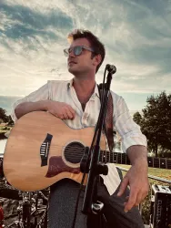 Ein Musiker sitzt mit einer Akustikgitarre und schaut in die Ferne. Im Hintergrund sind Bäume und ein schöner Himmel zu sehen.