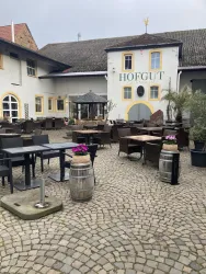 Ein gemütlicher Außenbereich eines Restaurants mit Tischen und Stühlen. Im Hintergrund befindet sich ein traditionelles Gebäude mit der Aufschrift "HOFGUT".
