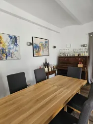 Ein moderner Essbereich mit einem langen Holztisch und Stühlen. An der Wand hängen Kunstwerke, und im Hintergrund steht ein Holzschrank mit Glaswaren.