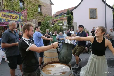 Eine fröhliche Stadtfest-Szene mit Menschen, die an einem Wettkampf teilnehmen. Auf dem Bild sind mehrere Teilnehmer zu sehen, die mit Eimern jonglieren, während eine begeisterte Menge zuschaut.