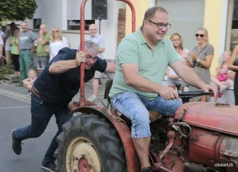 Zwei Männer auf einem alten Traktor, während eine Person den Traktor schiebt. Eine Menge Zuschauer feuert sie an.
