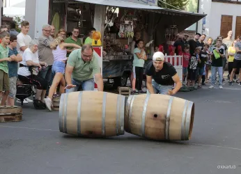 Zwei Männer rollen Holzfässer auf einer Straße, während sich eine Menschenmenge um sie versammelt hat. Es scheint ein Wettkampf oder ein Fest zu sein.