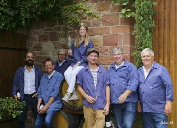 Eine Gruppe von sieben Personen steht vor einer Steinmauer, umgeben von Weinfässern. Alle tragen blaue Arbeitskleidung und lächeln freundlich in die Kamera.