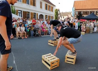 Zwei Männer springen auf Holzpaletten in einer Straßenveranstaltung. Eine große Menschenmenge schaut zu und feuert die Teilnehmer an.