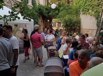 Eine belebte Gasse mit vielen Menschen, die an Tischen sitzen und sich unterhalten. Umgeben von grünen Pflanzen und gemütlicher Beleuchtung.