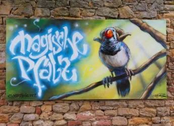 Ein farbenfrohes Wandgemälde eines Vogels auf einem Ast. Darunter steht der Schriftzug „magische Plätze“ in blauen Buchstaben.