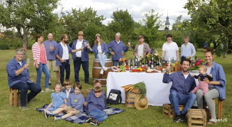 Eine Gruppe von Menschen feiert im Freien mit Essen und Getränken. Im Hintergrund stehen Obstbäume und ein schön gedeckter Tisch.