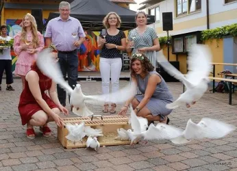 Eine feierliche Veranstaltung im Freien mit mehreren Personen. Weiße Tauben fliegen aus einem Käfig, während Gäste aufmerksam zuschauen.