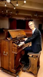 Ein Mann sitzt an einer alten Orgel und spielt. Der Raum hat eine elegante Atmosphäre mit warmen Licht.