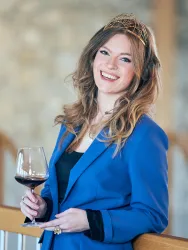 Eine lächelnde Frau in einem blauen Anzug hält ein Weinglas in der Hand. Sie trägt eine Krone und steht in einem eleganten Ambiente.