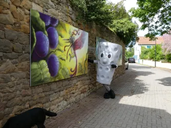 Eine Person in einem lustigen Kostüm steht an einer Steinmauer, neben einem bunten Kunstwerk mit Trauben. Im Hintergrund sind grüne Bäume und ein helles Wohnhaus zu sehen.