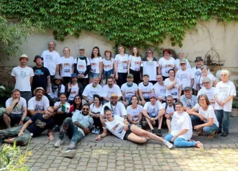 Eine große Gruppe von Menschen steht und sitzt zusammen in einem Innenhof. Viele tragen weiße T-Shirts und lächeln in die Kamera.