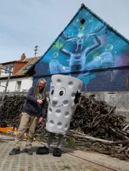Ein Mann steht neben einer großen, bulrigen Maskottchenfigur in Form eines Bechers mit Löchern. Im Hintergrund ist ein bunter Wandmalerei mit mystischen Motiven zu sehen.