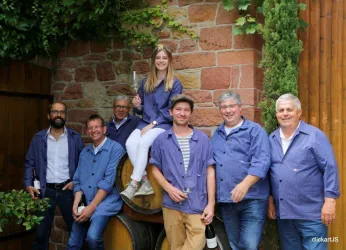 Eine Gruppe von sieben Personen posiert vor Weinfässern in einem Garten. Sie tragen blaue Arbeitskleidung und lächeln freundlich in die Kamera.
