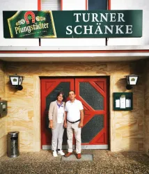 Ein Paar steht vor dem Eingang zur Turner Schänke, einem Gasthaus. Über der Eingangstür hängt ein Schild mit dem Namen des Lokals und dem Logo der Pfungstädter Brauerei.