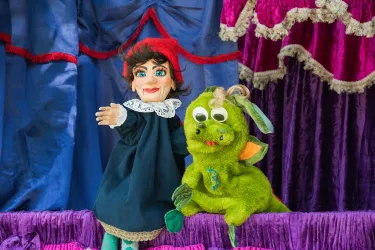 Eine Marionette mit roter Mütze und blauem Kleid steht neben einem grünen Drachen. Im Hintergrund sind bunte Vorhänge zu sehen.