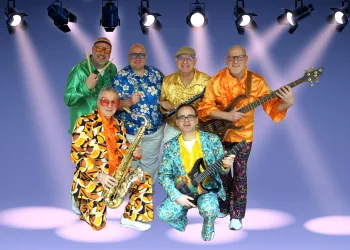 Eine bunt gekleidete Band posiert lächelnd auf einer Bühne. Im Hintergrund strahlen Scheinwerfer auf die Musiker.