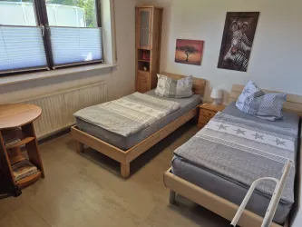 Ein einfaches Schlafzimmer mit zwei Einzelbetten und hellen Möbeln. Die Wände sind mit einem Bild und einem Regal dekoriert.