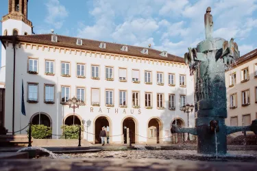 Ein historisches Rathaus mit mehreren Fenstern und einer großen Eingangstür. Im Vordergrund steht ein Brunnen mit skulpturalen Elementen.