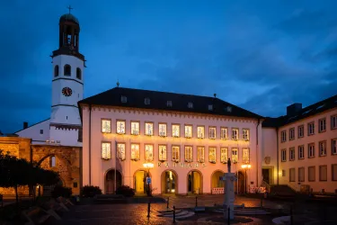 Ein historisches Gebäude bei Nacht, beleuchtet von warmem Licht. Im Hintergrund ist ein hoher Kirchturm zu sehen, umgeben von einem bewölkten Himmel.