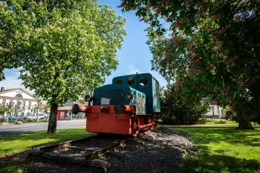 Eine alte Lokomotive steht auf Schienen, umgeben von grünen Bäumen. Im Hintergrund sind einige Gebäude sichtbar.