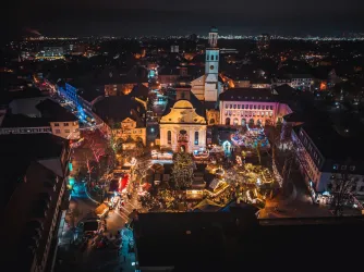 Ein festlicher Weihnachtsmarkt bei Nacht mit bunten Lichtern und einer beleuchteten Kirche. Es gibt zahlreiche Stände und eine große Weihnachtsbaummitte im Marktplatz.