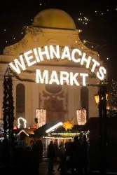 Ein festlich dekorierter Weihnachtsmarkt in der Nacht. Über dem Eingang leuchtet ein großes Schild mit der Aufschrift "WEIHNACHTSMARKT".