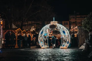 Ein festlich beleuchteter Weihnachtsmarkt mit einem großen, dekorativen Lichterschmuck. Menschen genießen die Atmosphäre und schlendern durch die Stände.