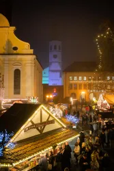 Ein festlicher Weihnachtsmarkt bei Nacht, umgeben von stimmungsvoller Beleuchtung. Im Hintergrund steht ein Kirchturm und es sind viele Menschen zu sehen.
