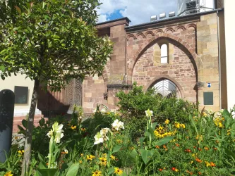 Eine historische Mauer mit einem großen, geschwungenen Fenster und bunten Blumen im Vordergrund. Die Szenerie ist grün und einladend, mit blauem Himmel im Hintergrund.
