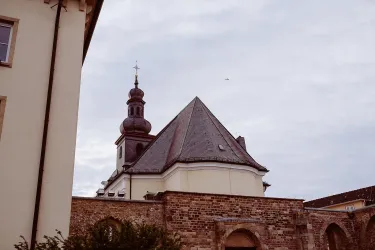 Ein historisches Gebäude mit einem spitzdachigen Kirchturm. Der Himmel ist bewölkt und die Architektur zeigt eine Mischung aus Steinen und modernem Design.