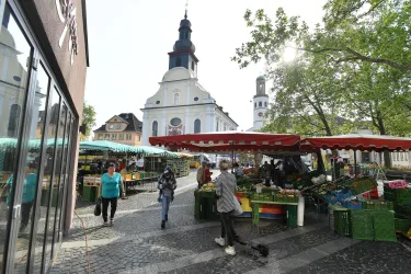Ein lebhafter Wochenmarkt mit bunten Ständen und frischem Obst und Gemüse. Im Hintergrund ist eine Kirche mit Kuppelturm zu sehen.