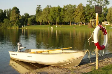 Ein kleiner Boot am Ufer eines ruhigen Sees. Im Hintergrund sind grüne Bäume und Menschen, die die Natur genießen.