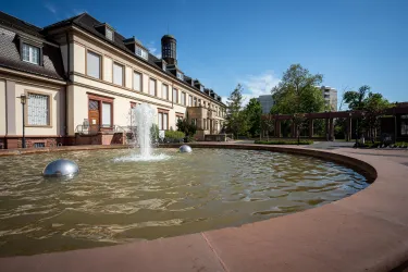 Ein schöner Brunnen mit Wasser im Vordergrund vor einem eleganten Gebäude. Im Hintergrund sind grüne Bäume und ein klarer blauer Himmel zu sehen.