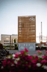 Ein modernes Holzgebäude mit der Aufschrift "Frankenthal" und einem Fahrrad-Symbol. Im Vordergrund sind Blumen und Fahrräder sichtbar.