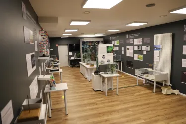 Ein moderner Ausstellungsraum mit Tischen, auf denen verschiedene Modelle und Informationsmaterialien präsentiert werden. Die Wände sind mit Plakaten und Erklärungen zum Thema ausgestattet.