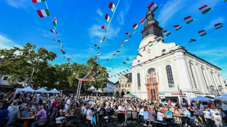 Ein belebter Platz mit vielen Menschen, die an einem Fest teilnehmen. Bunte Fahnen wehen über der Szene und eine Kirche ist im Hintergrund sichtbar.