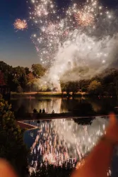 Ein beeindruckendes Feuerwerk über einem See, das das Wasser zum Glitzern bringt. Zuschauer genießen das Spektakel am Ufer.
